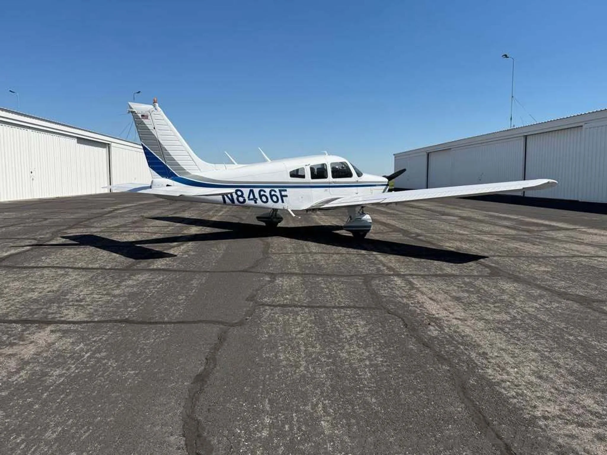 A white Piper Warrior II aircraft with blue and gray stripes and registration N8466F parked on an asphalt apron.