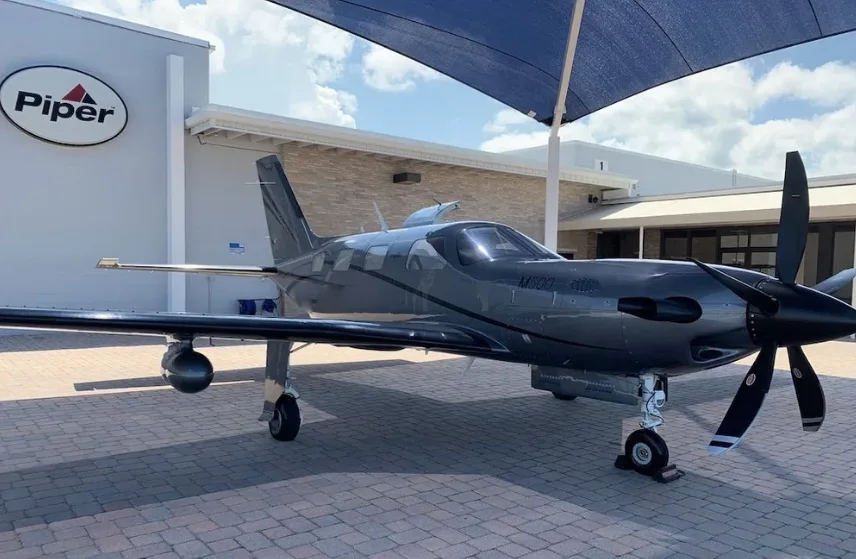 A dark grey Piper M600 SLS aircraft is parked on a paved area next to a building with a Piper logo.