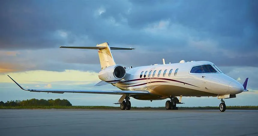 A white private jet with a dark stripe, parked on an airfield under a dramatic sky.