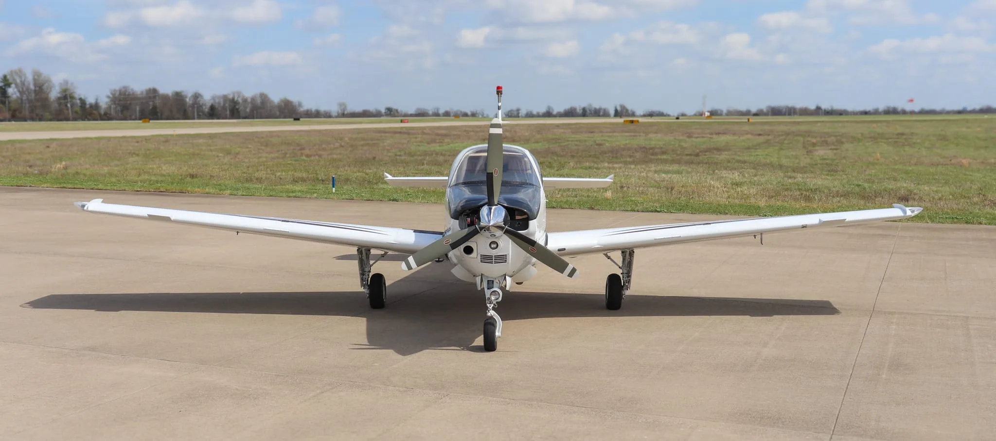 A head-on view of a white Beechcraft Bonanza G36 aircraft with a black nose, parked on an airfield with a grassy background.