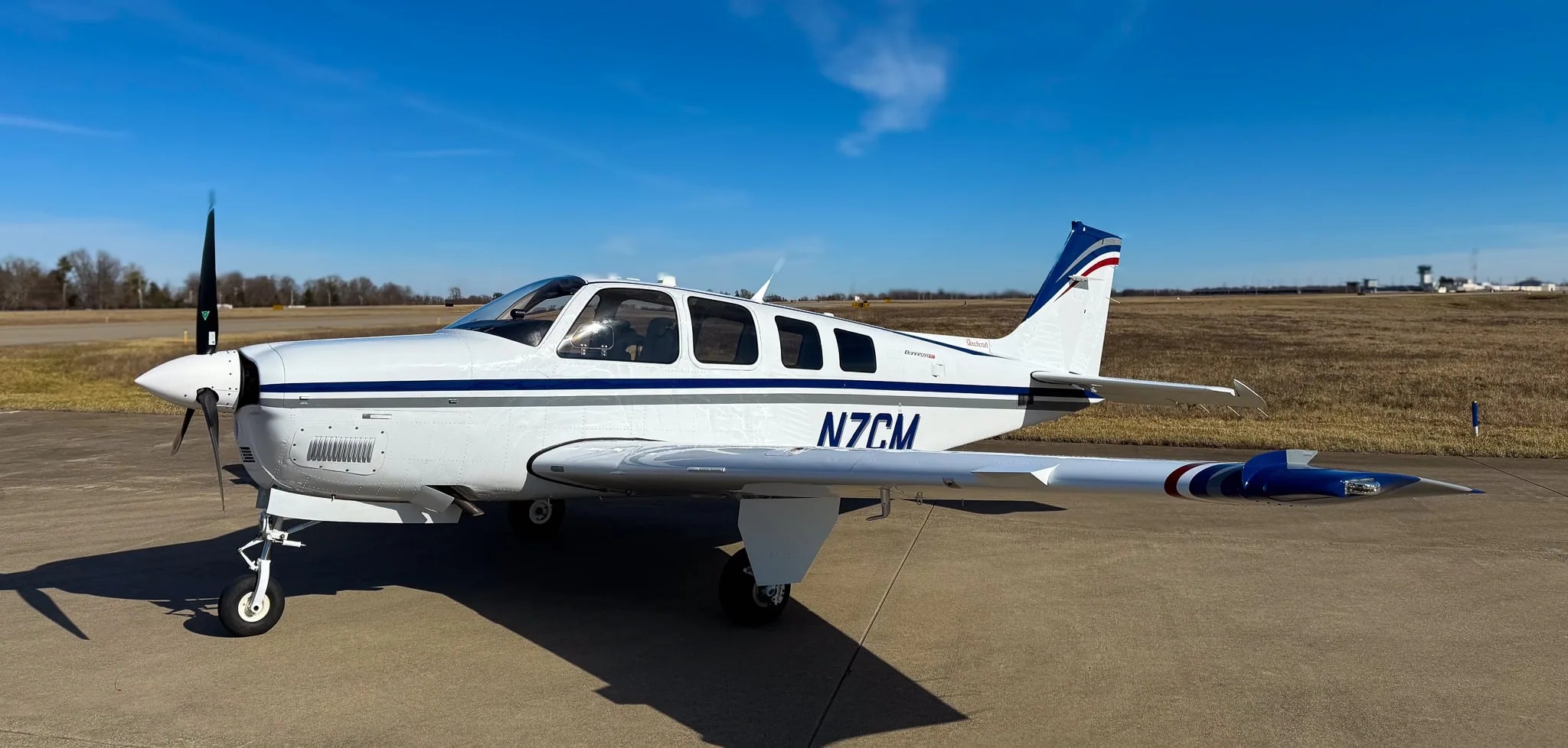 A white Beechcraft Bonanza G36 with blue and grey stripes, registration N7CM, parked on an airfield under a clear blue sky.