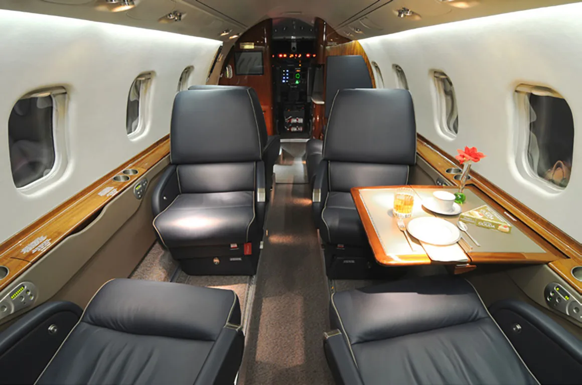The luxurious interior cabin of a private jet features black leather seats, wood trim, and a table set with drinks and a red flower.
