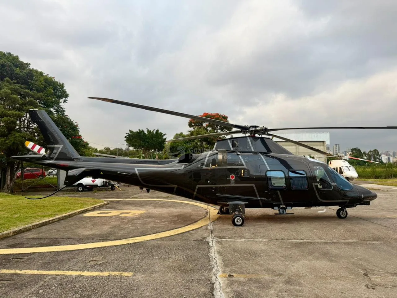 A black AgustaWestland AW109 helicopter with a white stripe on its tail, parked on an apron.