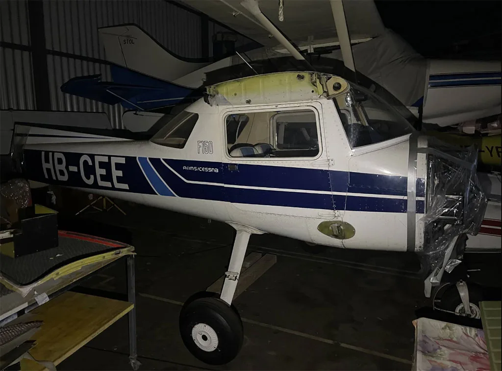 A white and blue Cessna F150 aircraft, registration HB-CEE, with its engine cowling removed, sits in a dimly lit hangar.