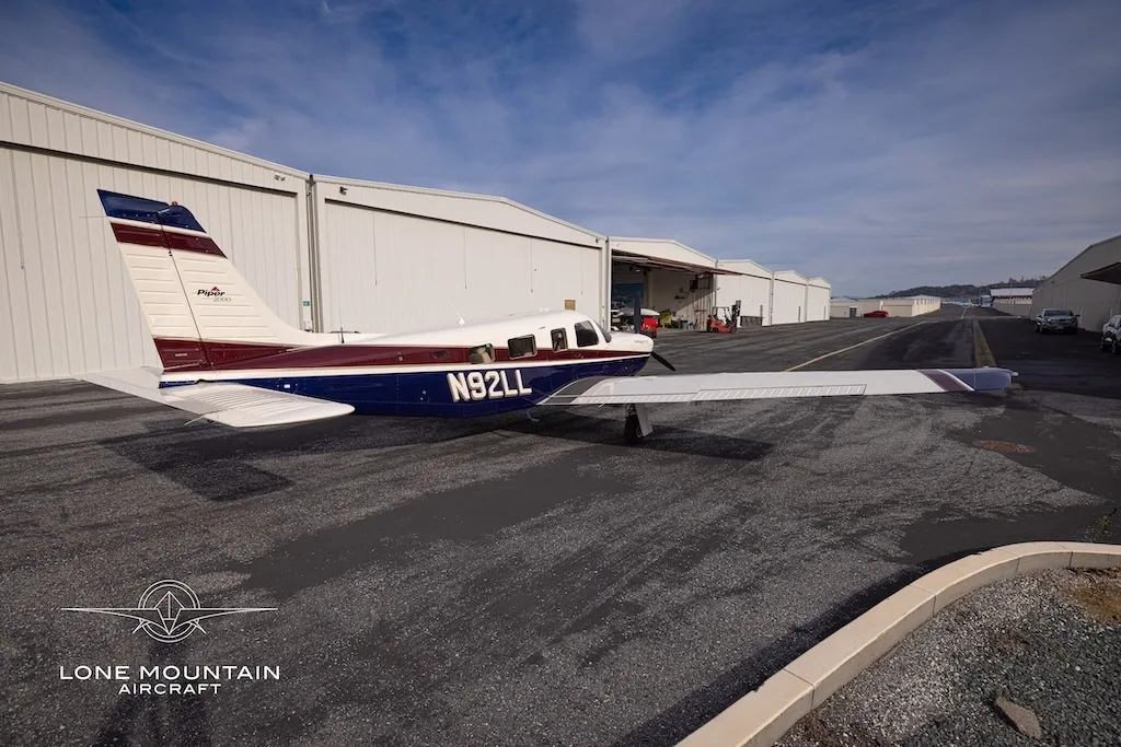 A rear three-quarter view of the Piper Saratoga II TC aircraft (N92LL) with blue and white paint scheme parked on an asphalt apron next to hangars.