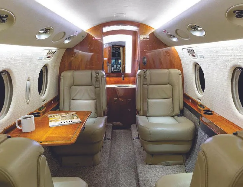 View of the aircraft cabin interior with tan leather seats, a wooden table with a mug and magazine, and wood paneling.