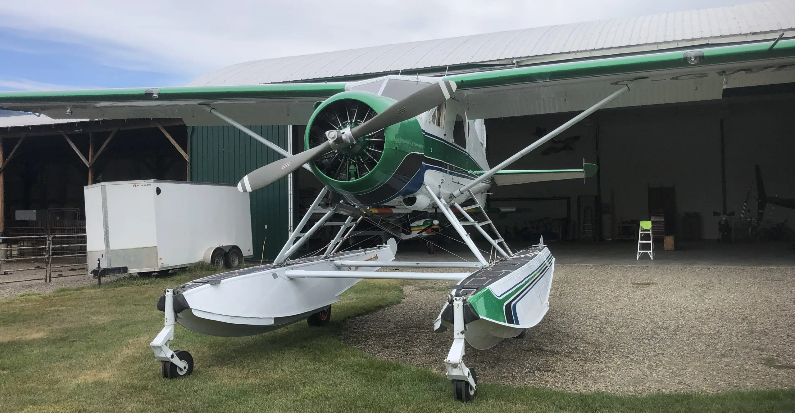 A green, white, and blue single-engine floatplane with retractable landing gear on its floats, parked on gravel in front of a hangar.