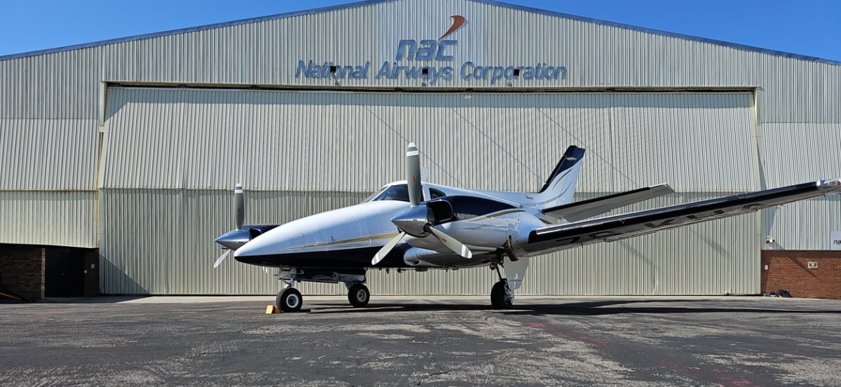 A white and black twin-engine aircraft with propellers parked on an asphalt apron in front of a large hangar.