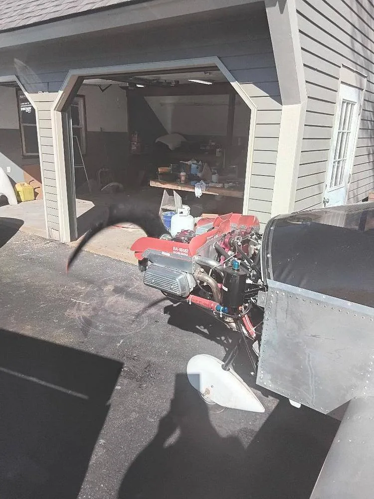 A partially assembled light aircraft with its red engine exposed, parked on a driveway next to a garage.