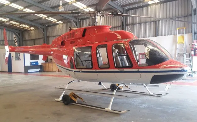 A red and white helicopter with skids is parked inside a hangar, viewed from the front-right.