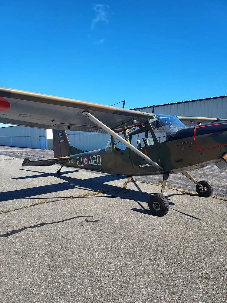 A side view of a single-engine, high-wing aircraft with olive drab paint and red wing markings, parked on an airfield.