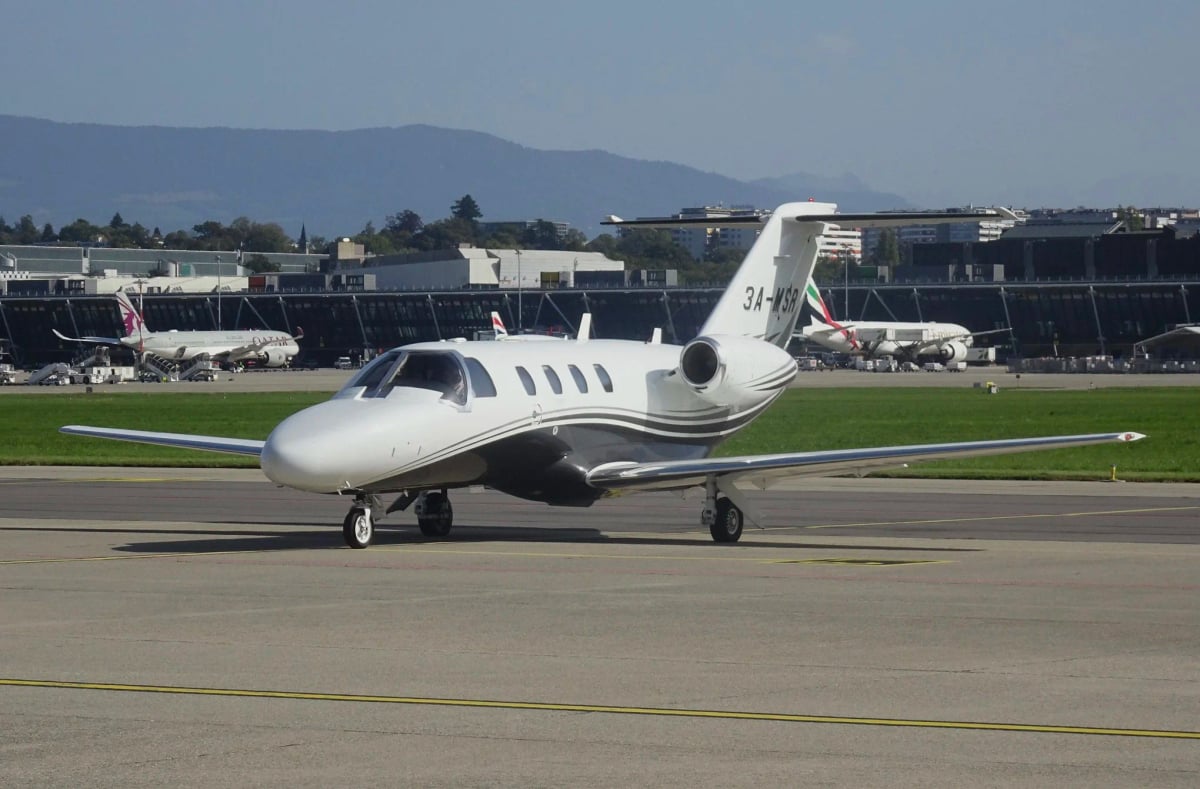 A white Cessna CitationJet with grey and black stripes, registration 3A-MSR, taxiing on an airport tarmac with other aircraft in the background.