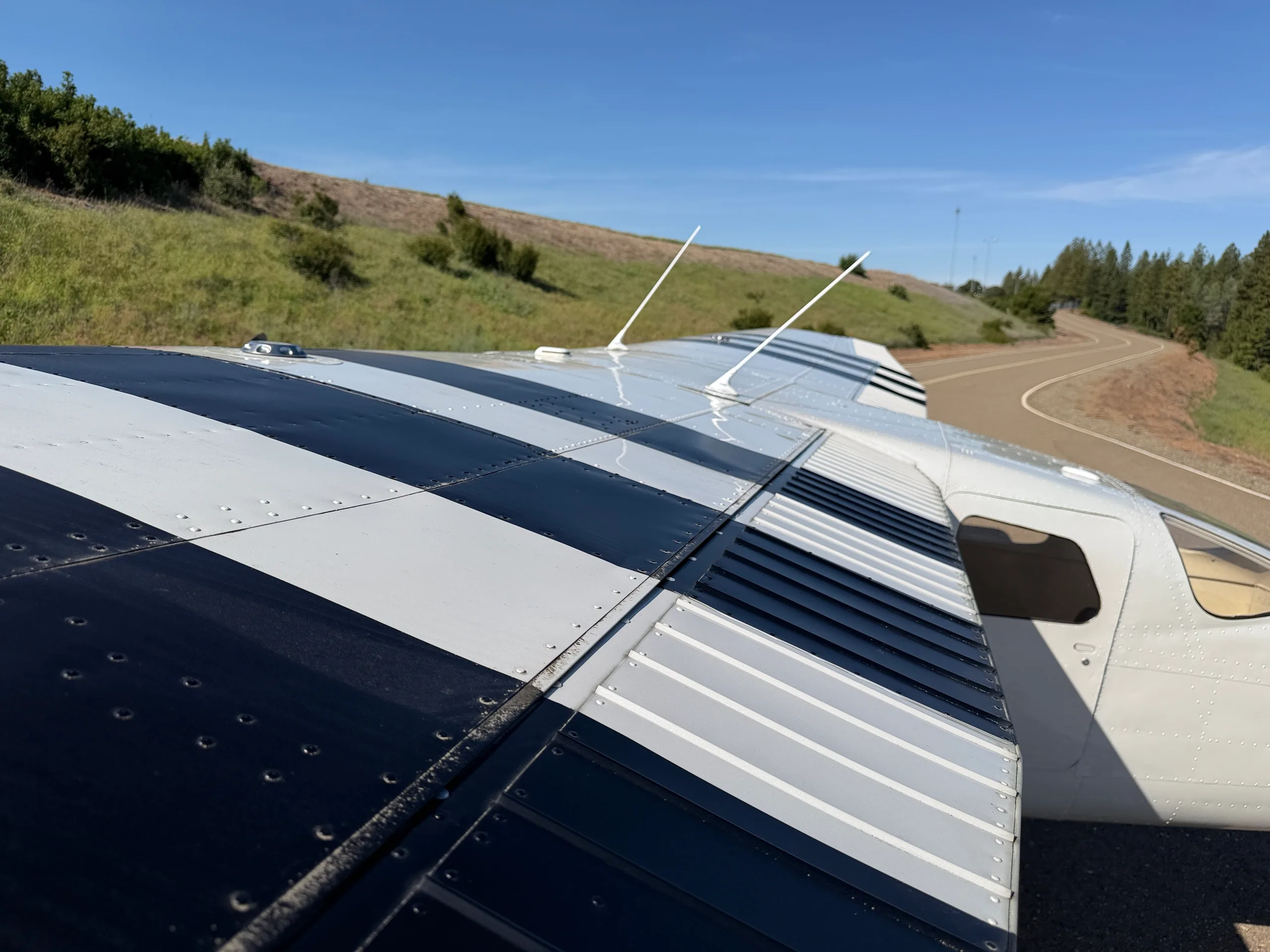Top-down view of the black and white striped wing of a Cessna 206, with a road and green hills in the background.