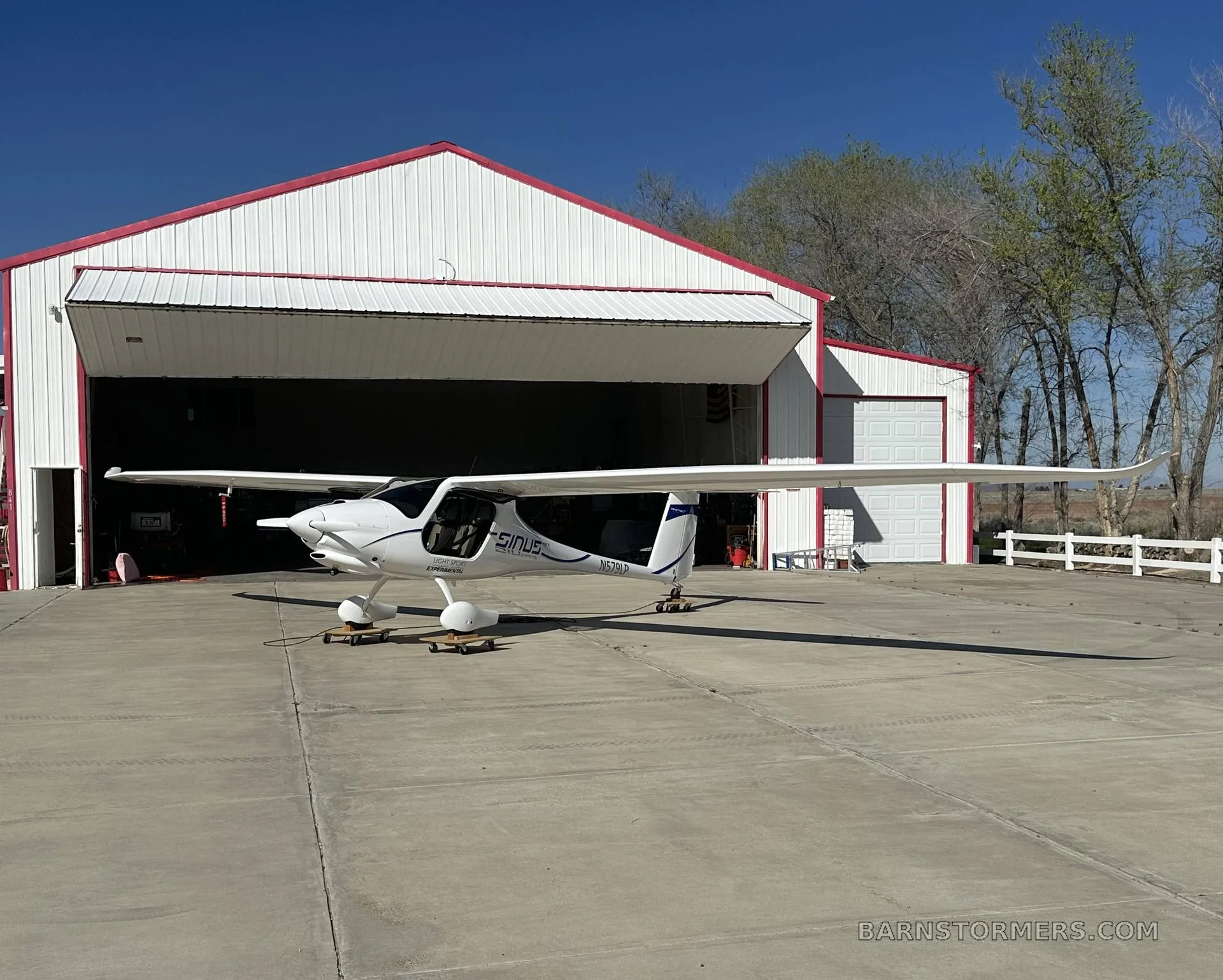 A white Pipistrel Sinus aircraft with blue accents is parked on an apron in front of a hangar on a sunny day.