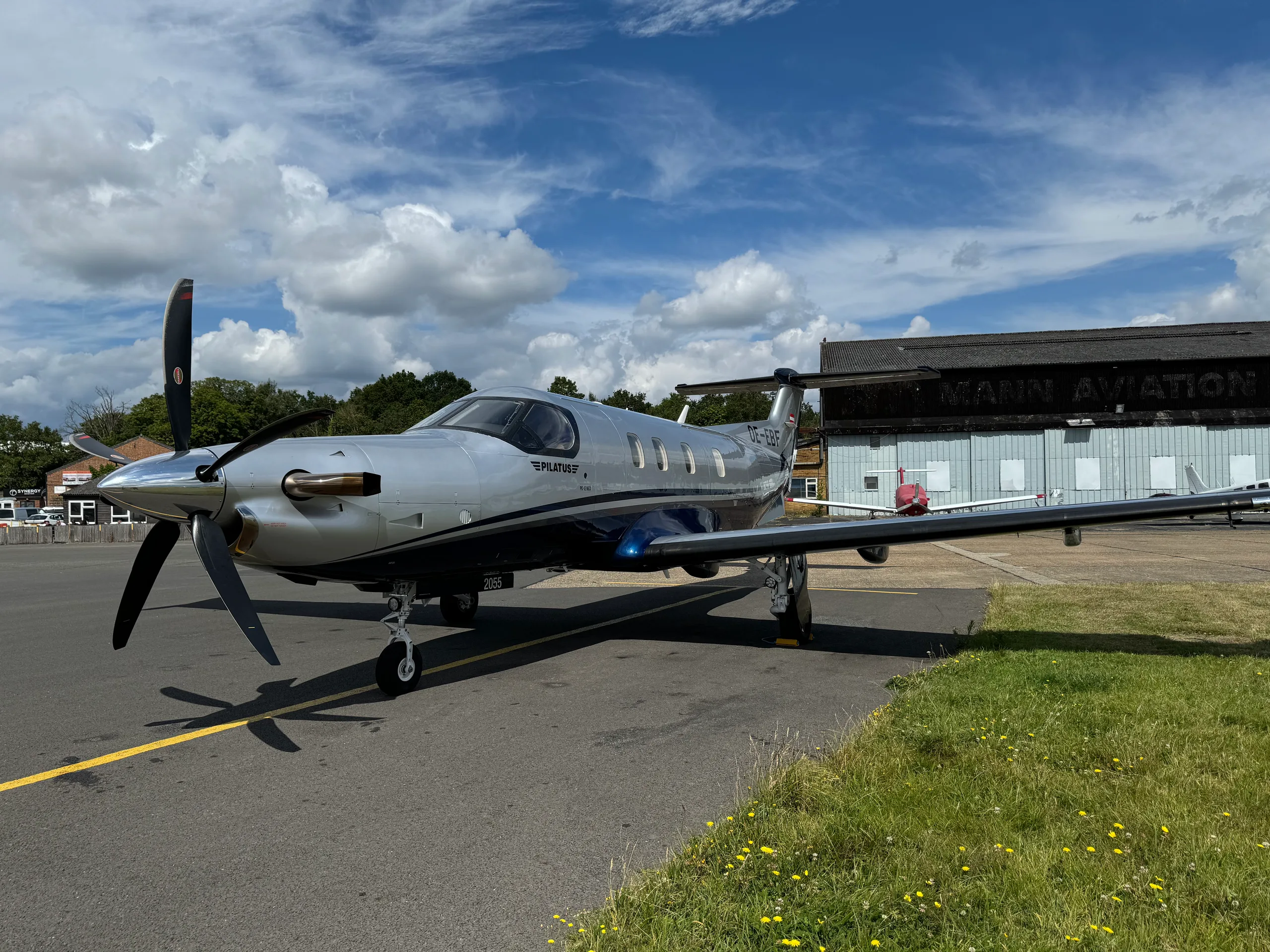 A silver and blue Pilatus PC-12 NGX turboprop aircraft with registration OE-EBF is parked on an airfield tarmac in front of a Mann Aviation hangar under a partly cloudy sky.