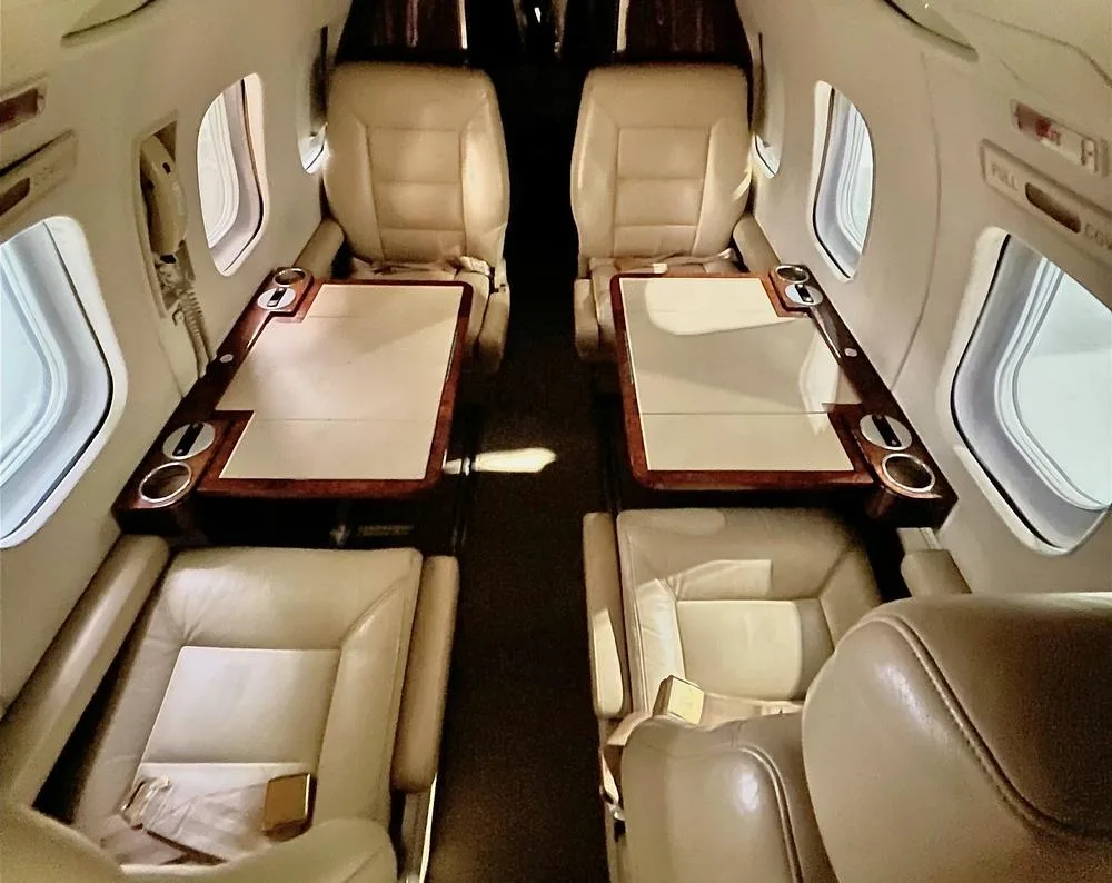 View of a private jet cabin with two sets of beige leather seats facing each other, each with a fold-out table.