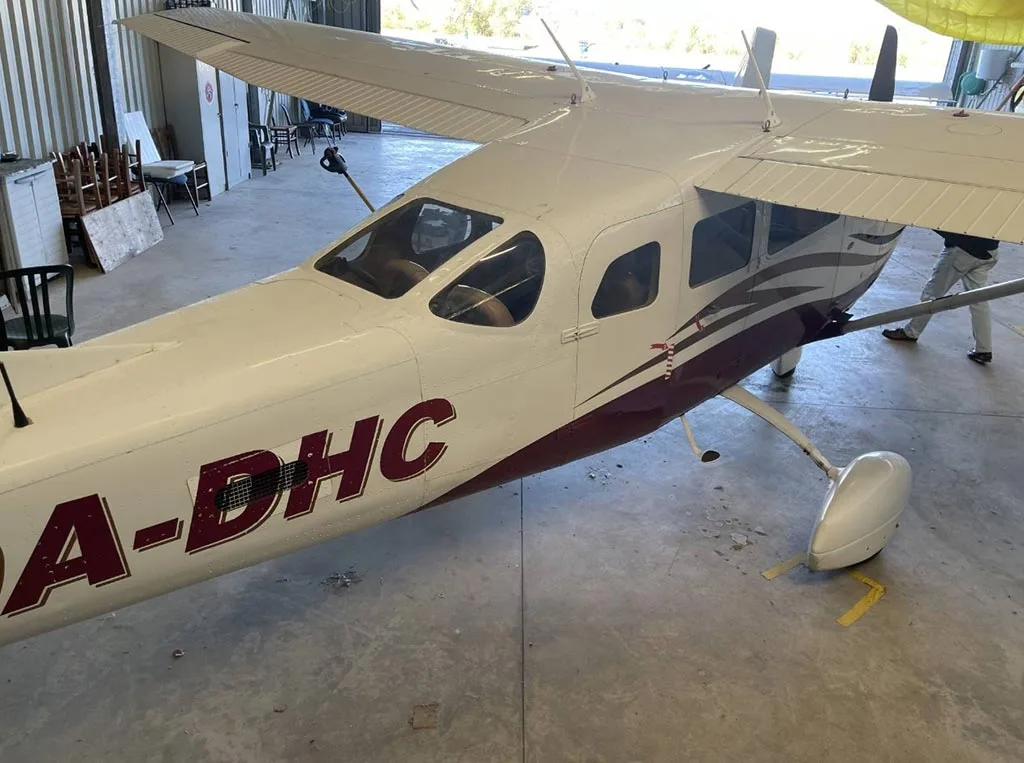 A rear-side view of the white and red Cessna 206/210 aircraft 9A-DHC, showing the fuselage and tail.