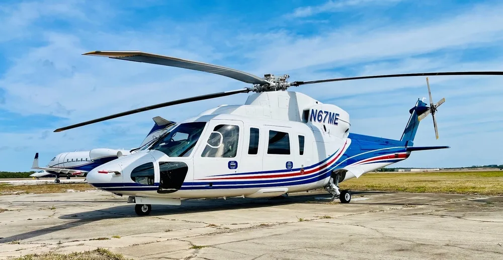 Front-right view of a white and blue helicopter (N67ME) with red stripes on an airfield under a blue sky.