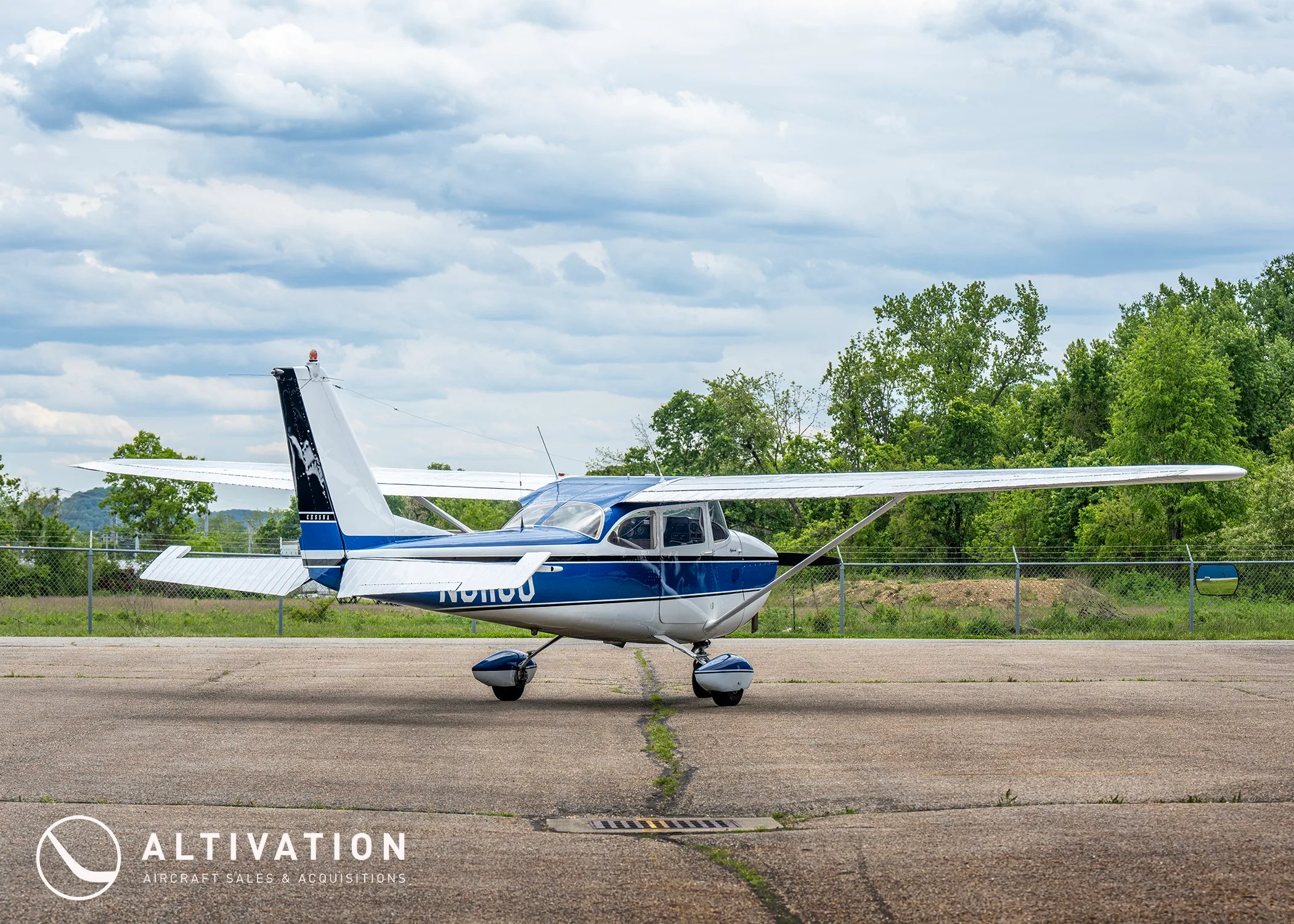A side view of the white and blue Cessna 172 Skyhawk N8118U parked on an asphalt ramp with trees and a fence in the background.