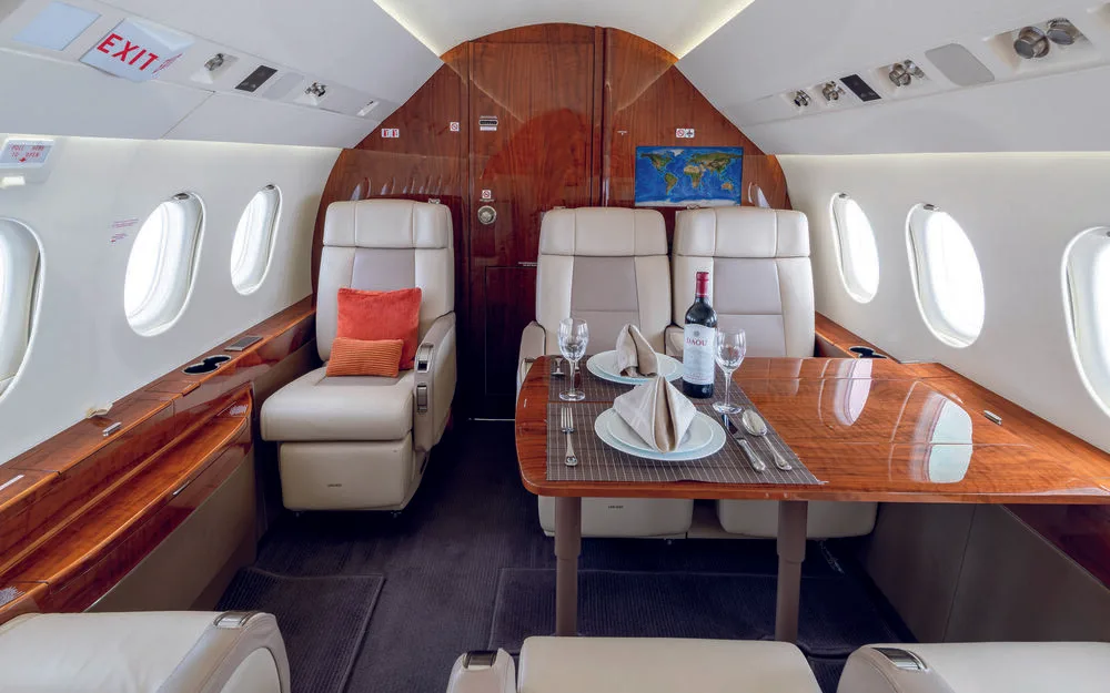 The elegant cabin interior of a business jet, showing beige seats, a wooden dining table with wine and place settings, and a world map display on the bulkhead.