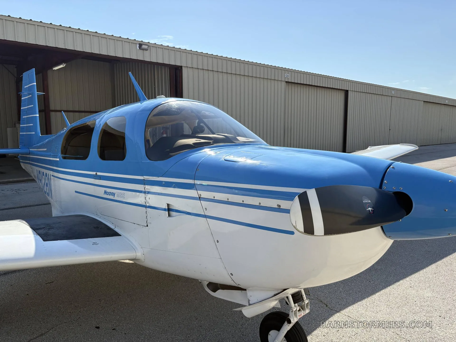 A blue and white Mooney M20 aircraft (N9128N) parked on an airfield with a hangar in the background, viewed from the front-left.