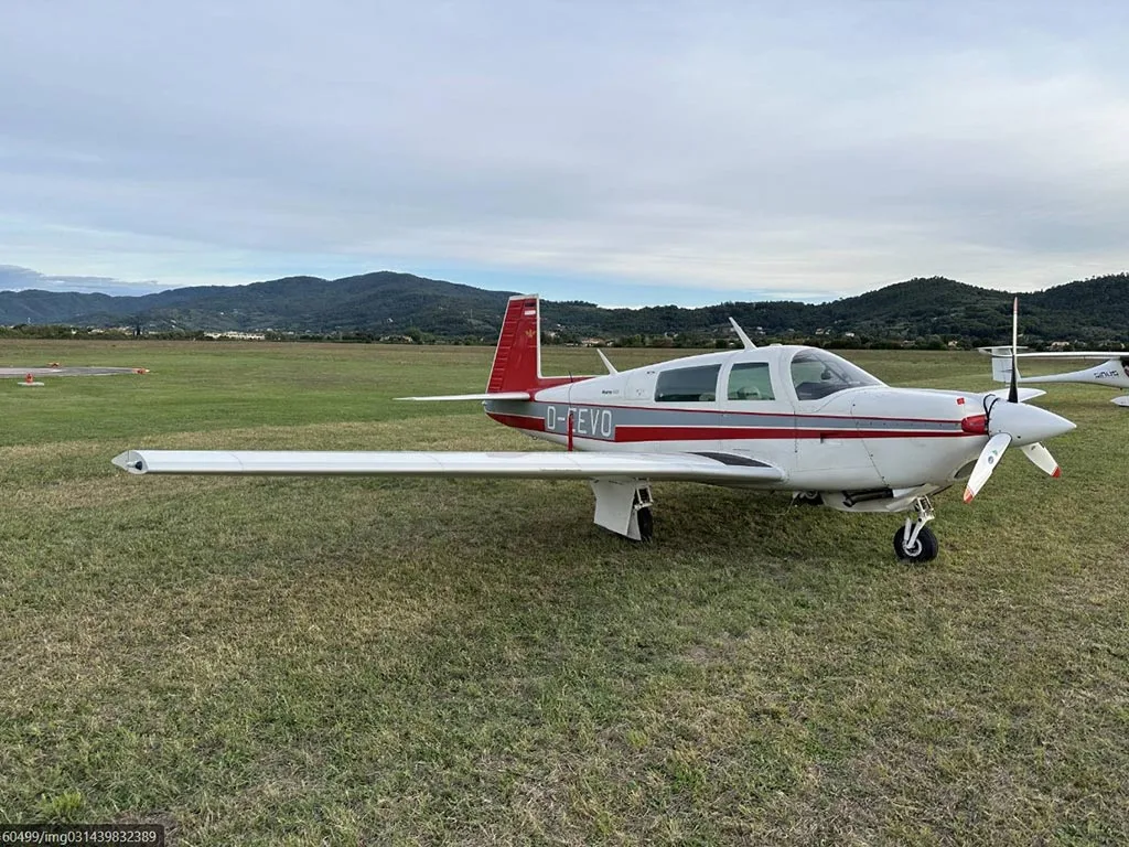 A white and red Mooney M20J aircraft (D-EEVO) parked on a grassy field with mountains in the background.