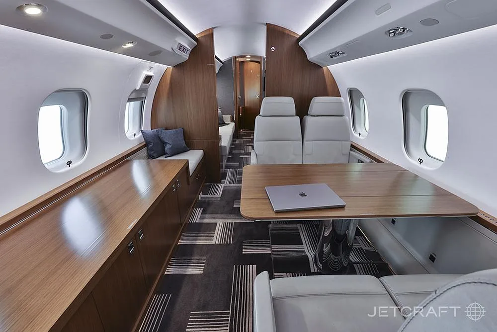 A view of the aircraft cabin showing a long wooden credenza, a table with a laptop, and light grey seats.
