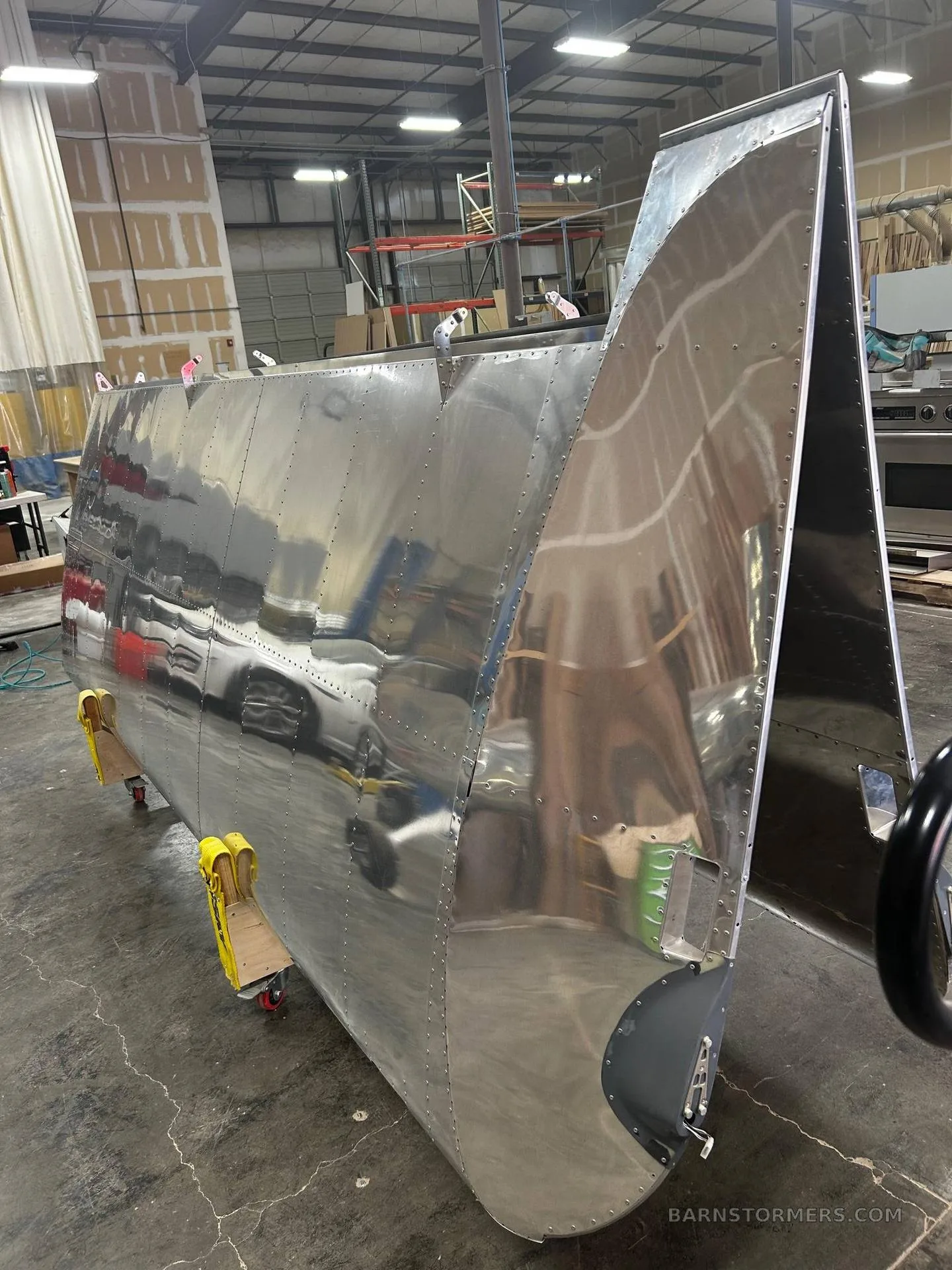 A large, polished aluminum aircraft fuselage section with rivets, resting on stands in a workshop.