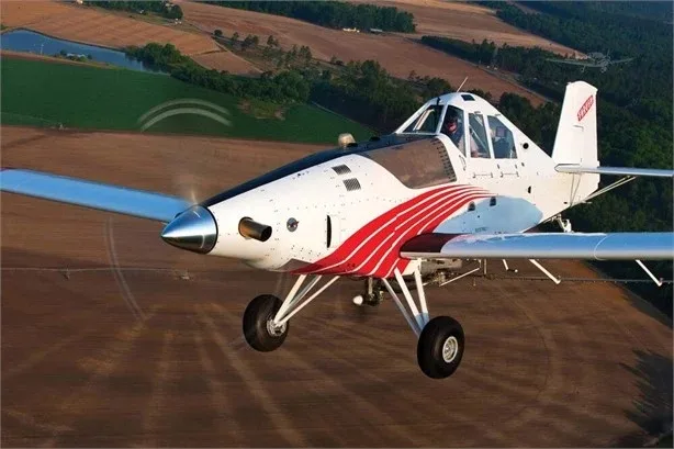 A white agricultural sprayer aircraft with red stripes flying low over brown fields.