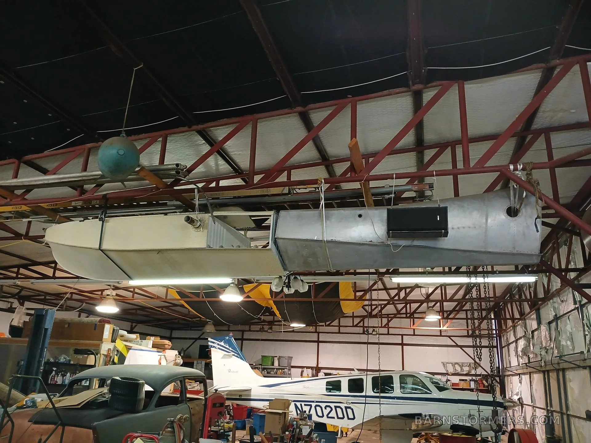 A large aircraft float or fuselage hangs from the ceiling above a white and blue Beechcraft Baron (N702DD) in a hangar.