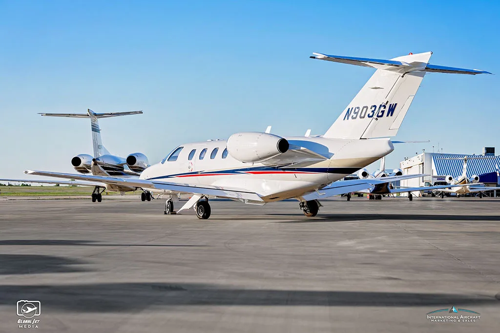 A rear view of a white Cessna Citation CJ3 (N903GW) with blue and red stripes parked on an airfield.
