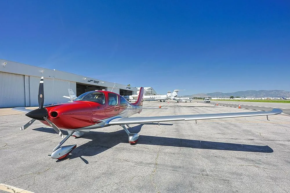 A red and silver Cirrus SR22 aircraft with registration N218JS parked on an airport tarmac with mountains in the distance.