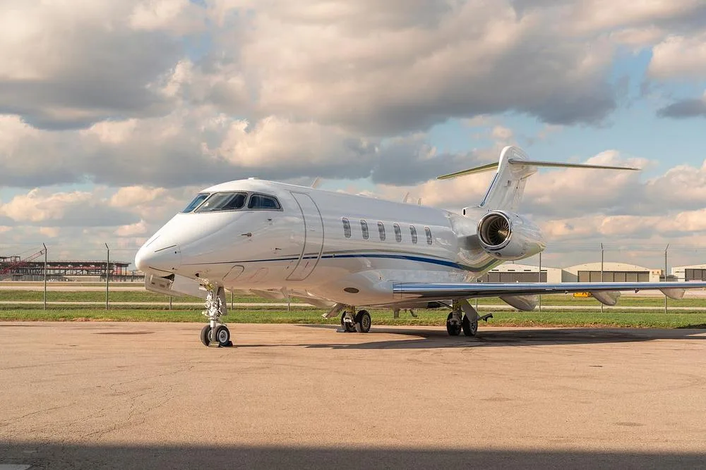 A white Bombardier Challenger 350 business jet with blue stripes parked on the tarmac, viewed from the front-side.