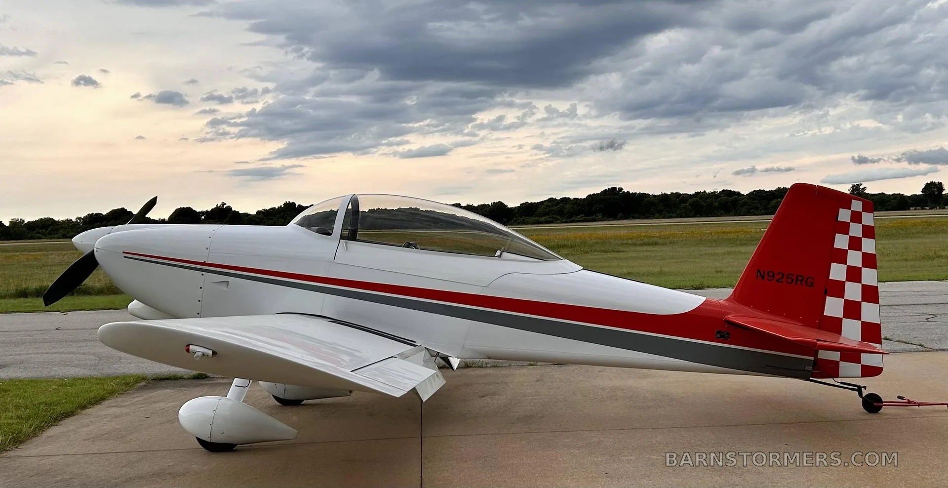 A white single-engine aircraft with red and gray stripes and a checkered red tail, parked on an airfield under a cloudy sky.