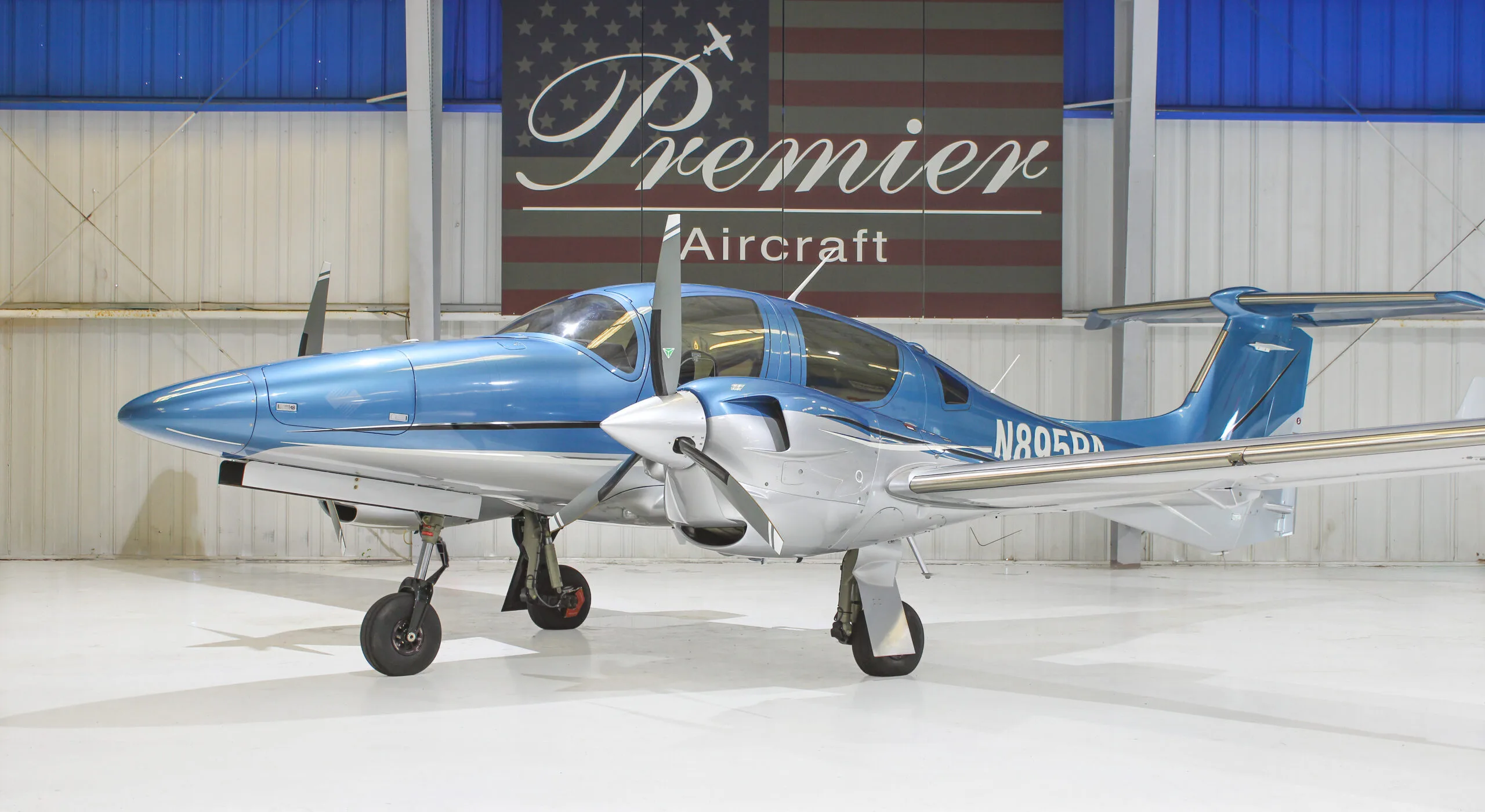 A blue and silver Diamond DA62 aircraft parked in a hangar with a 'Premier Aircraft' banner in the background.