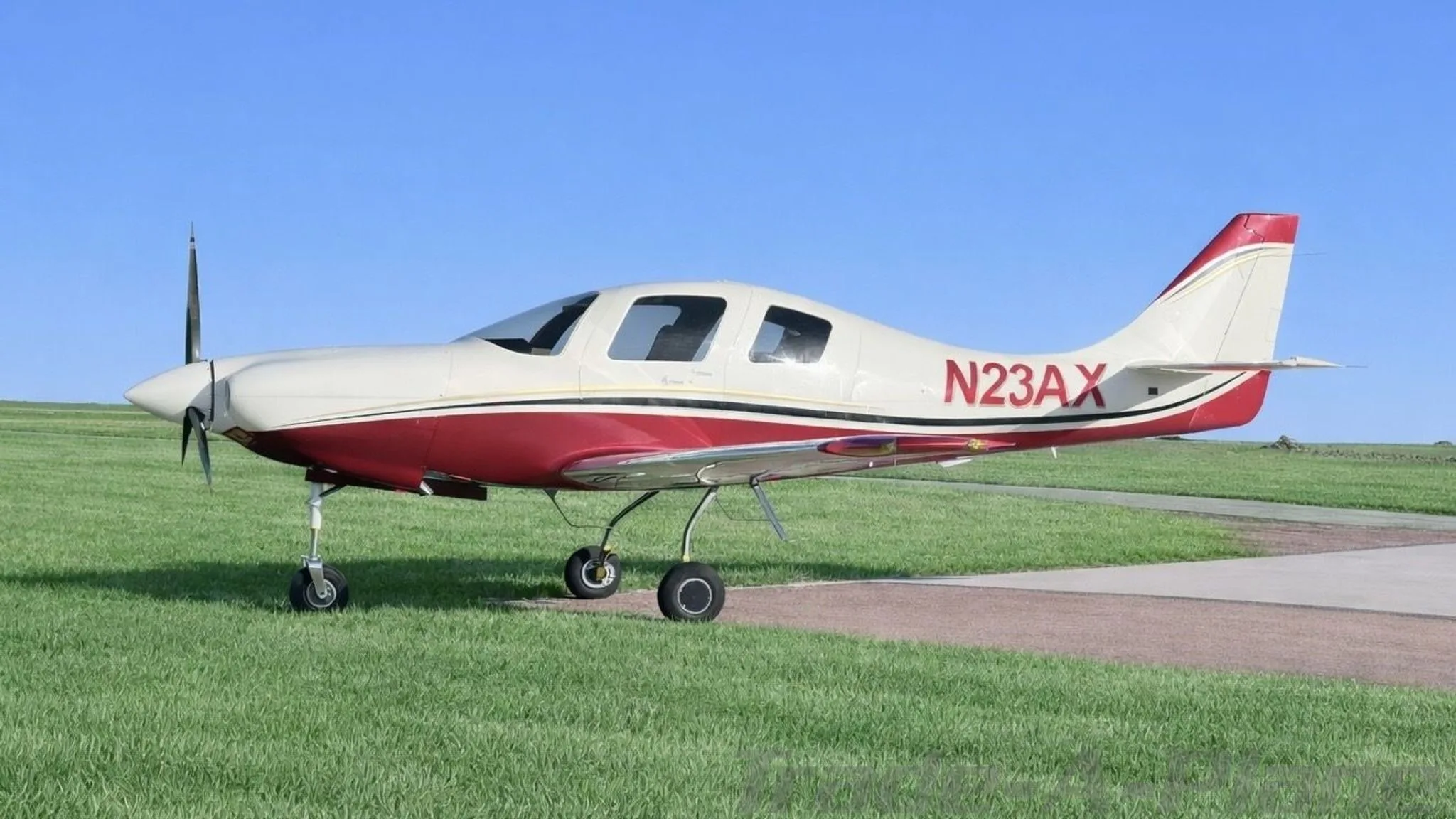 A white and red single-engine aircraft, N23AX, is parked on a grassy field next to a paved path under a clear blue sky.