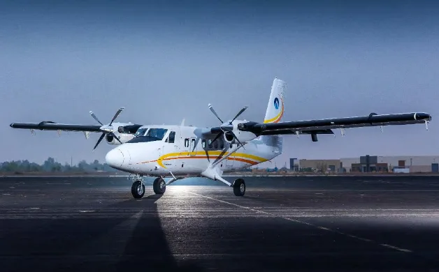 A white and yellow twin-engine turboprop aircraft, likely a DHC-6 Twin Otter, is parked on an asphalt runway under a bright sky.