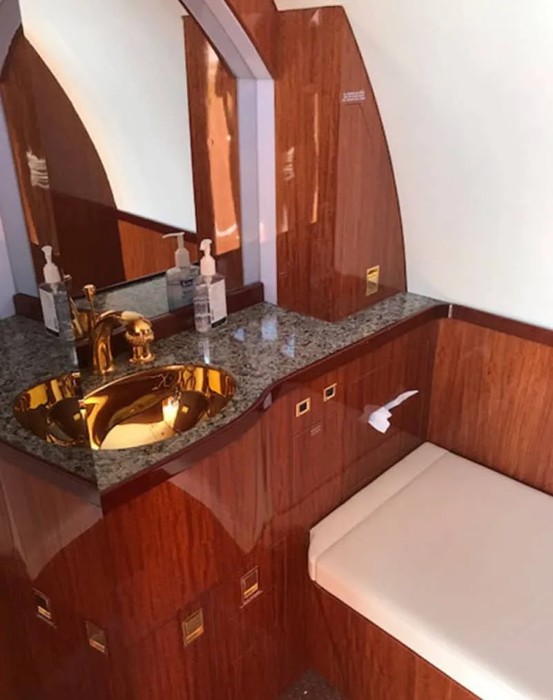 An aircraft lavatory features a gold sink, granite countertop, and polished wood paneling.