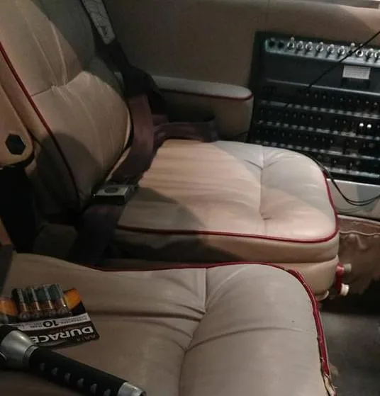 Close-up view of two beige leather aircraft seats with red piping and seatbelts, with equipment in the background.