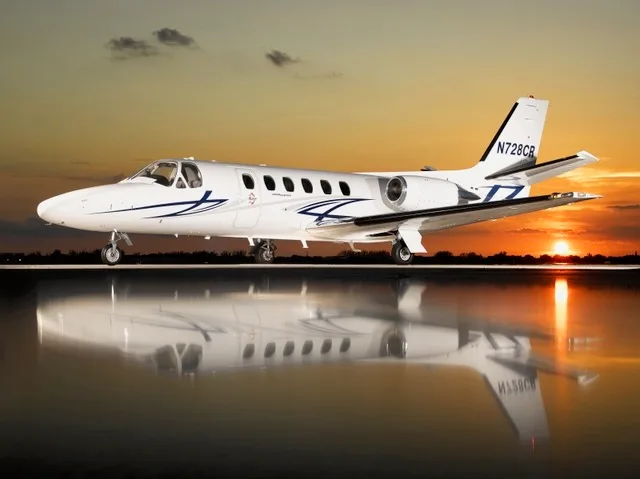 A white Cessna Citation business jet with blue and black stripes is parked on a tarmac, reflecting in a wet surface, against a vibrant sunset sky.