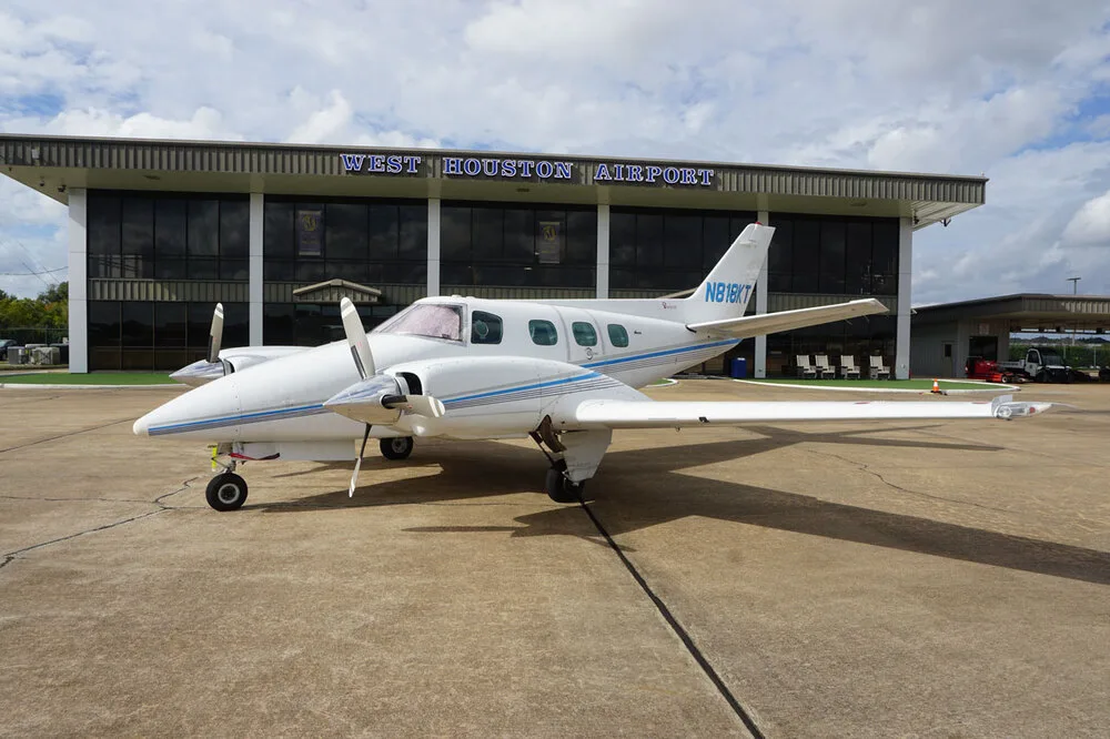 A white twin-engine propeller aircraft with blue stripes, registration N818KT, parked on the tarmac with the West Houston Airport building in the background.