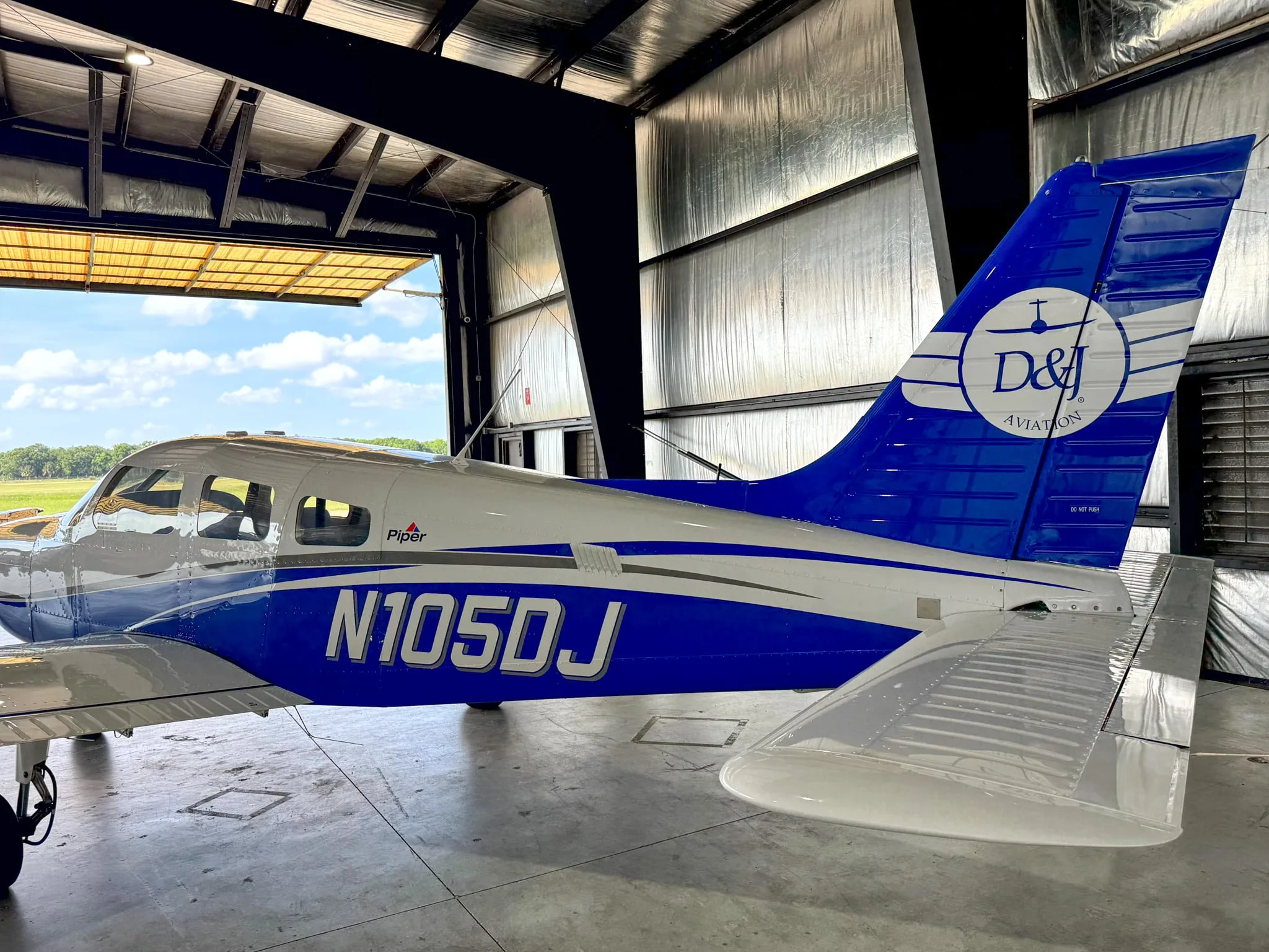 A side view of the white and blue Piper Archer TX, N105DJ, parked in a hangar with a view of the airfield outside.