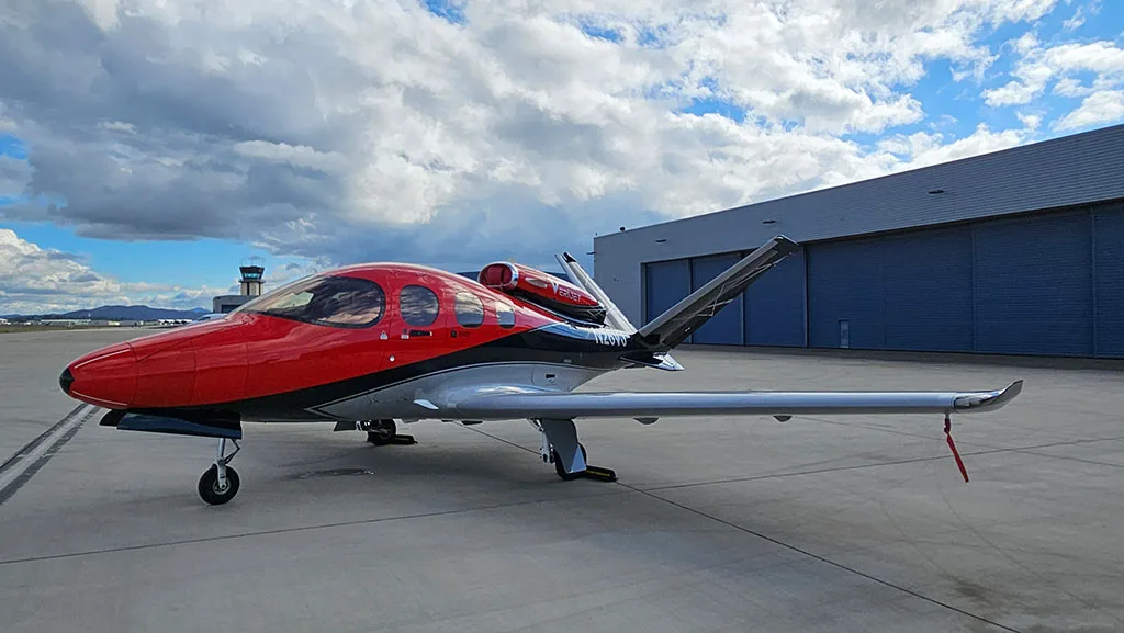 A side view of a red, black, and silver Cirrus Vision Jet (N26VJ) parked on an airport tarmac with an air traffic control tower in the distance.