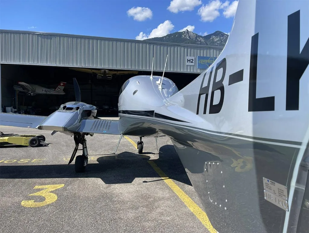 The tail section of a silver Diamond DA42 aircraft with registration HB-LY parked next to a hangar.
