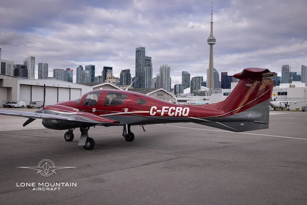A red and grey Diamond DA62 aircraft with registration C-FCRQ parked on an airfield, viewed from the side.