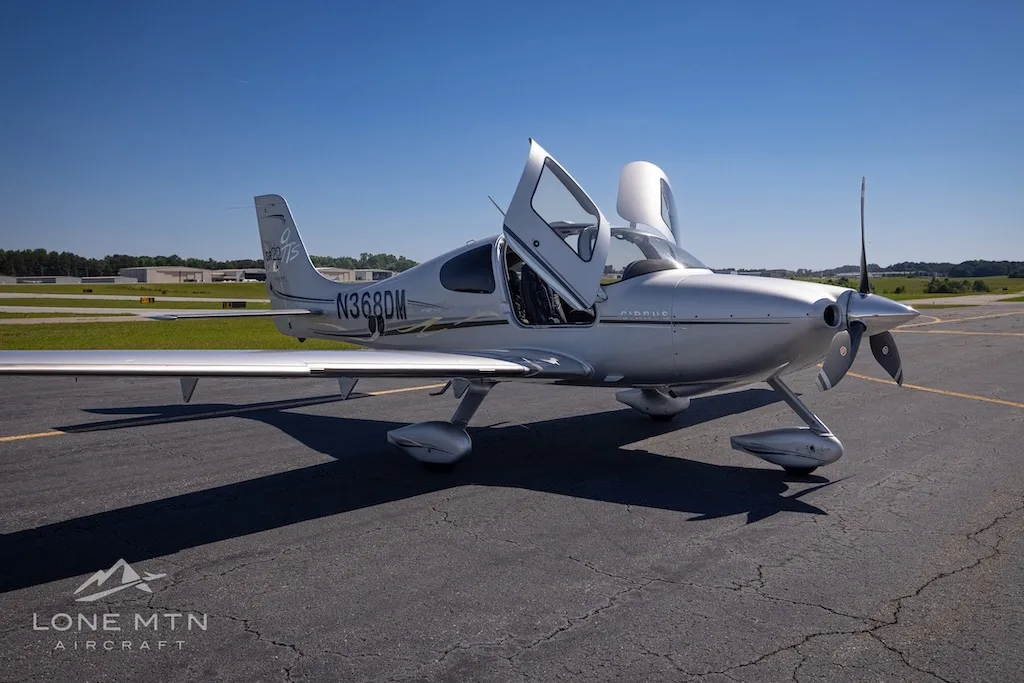 A silver Cirrus SR22 Turbo aircraft (N368DM) with its doors open, parked on an asphalt runway under a clear blue sky.