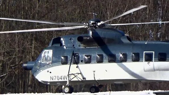 A large dark blue and white helicopter with registration N704WY is shown on a snowy ground with bare trees in the background.