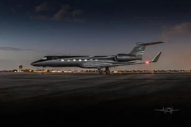 A dark grey Gulfstream G550 (N550AG) private jet with a light grey stripe on the tail, parked on an airport tarmac at dusk.