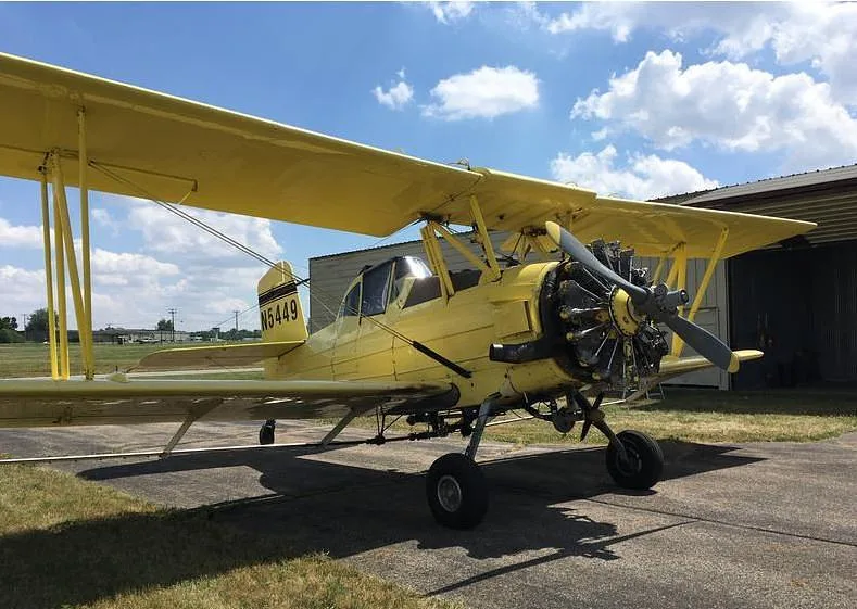 A yellow biplane crop duster, registration N5449, parked on an airfield with hangars in the background.