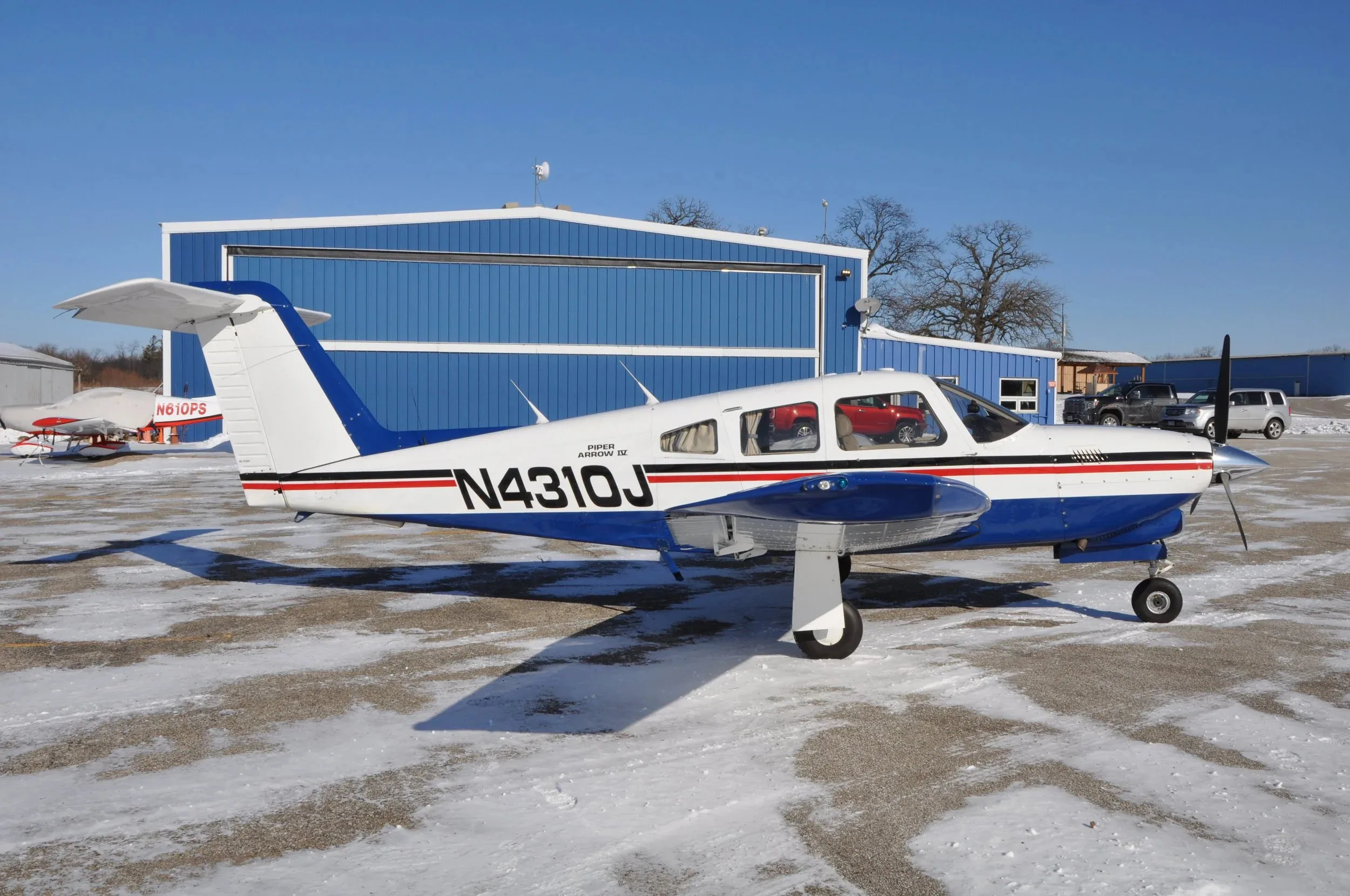 A white and blue Piper Arrow IV aircraft with registration N4310J parked on a snowy tarmac in front of a blue hangar.
